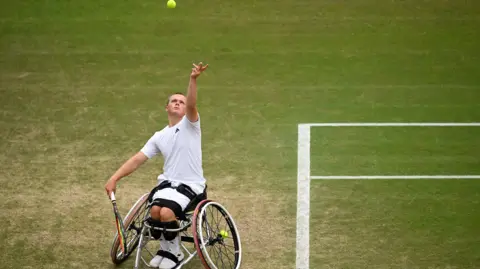 Getty Images Wheelchair tennis star Ben Bartram tosses the ball up to serve on a grass court