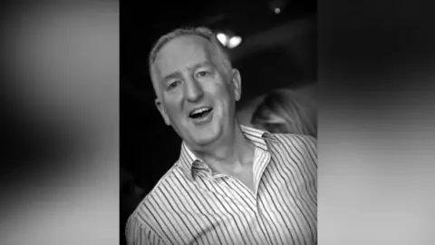 The black and white image shows Nick in a stripey shirt smiling. He has short grey hair and his top buttons are undone.