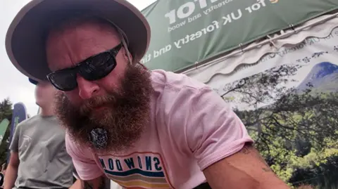 YouTube A man with a bushy beard, wearing a pink T shirt, looks at the camera