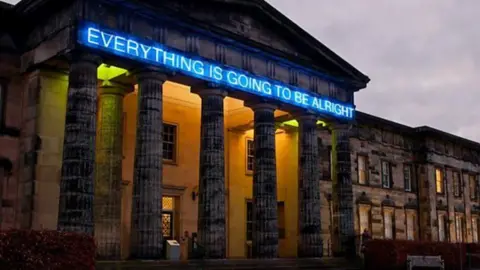 Harris Partnership A blue neon sign reading "everything is going to be alright" is installed on a large building with six pillars.