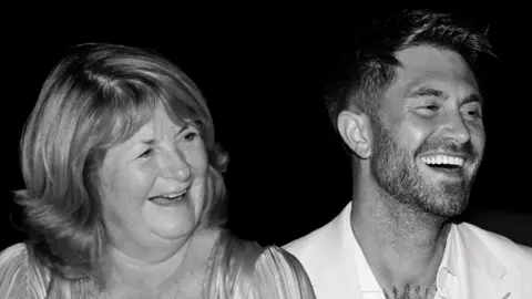 Ross Worswick and his mother Judith are sitting side by side and are laughing while staring off camera in this black and white photograph.