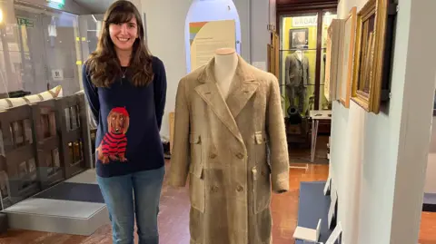 Curator Dayna Woolbright wears a blue sweatshirt with a Dachshund dog on the front. She is standing next to a pale cream coat which is spun from wool created from the shed hair of a St Bernard dog. 