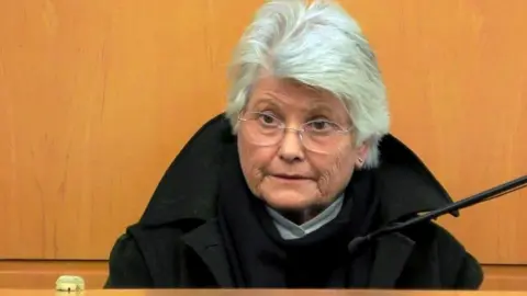Firecrest Films A 77-year-old woman with white hair and glasses, wearing a black coat, sits in the witness box at the High Court in Glasgow