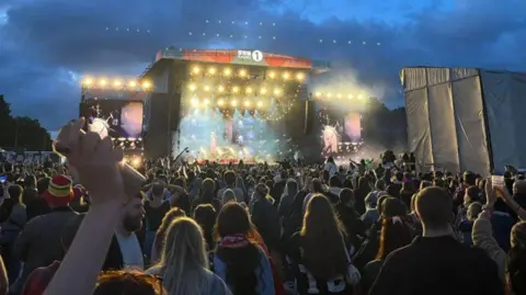 A view from the crowd looking towards a stage lit up . The crowd is in silhouette with some people holding their hands in the air