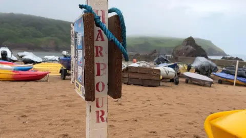 A post with a word Harbour on it and some rope on a beach with boats behind it.