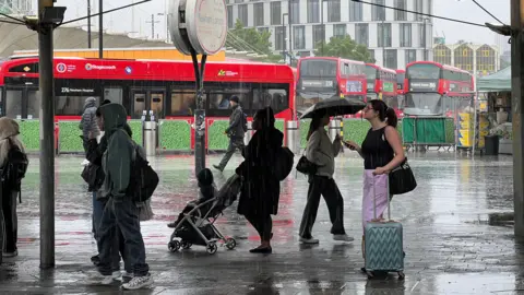 People stand under a shelter in Newham while it rains. There are buses in the background.