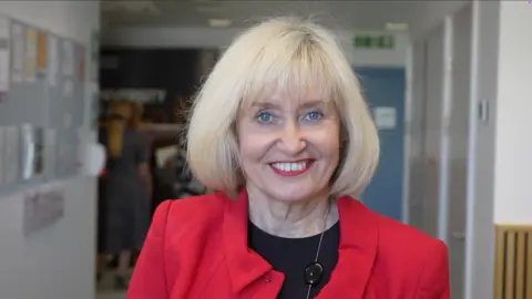 Jamie Niblock/BBC Professor Jenny Higham smiles at the camera. She has short blonde hair and wears a red blazer with a black top underneath.