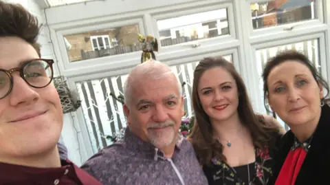 Family handout A selfie with four people in a row smiling at the camera. On the left is a man with brown hair and glasses wearing a burgundy shirt, then an older man with a grey beard and flowery purple shirt. On the right is a woman with long brown curly hair and red lipstick, and on the end is a woman with dark hair wearing a red dress.