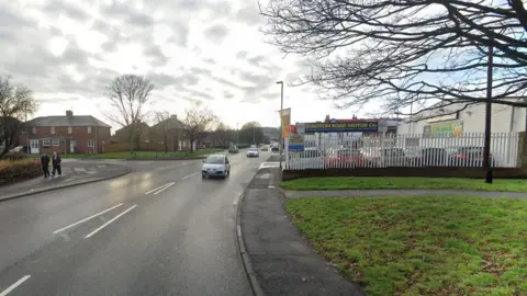 Dunston Road in Gateshead. A garage called Dunston Road Motor Co stands behind a fence to the right of the road. There is a patch of grass in front of the garage to the side of the pavement. Cars are travelling down the road towards the camera.