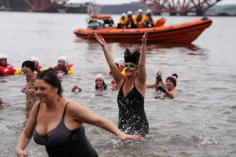 PA Media People in swimming costumes and hats race out of the water in the Forth of Firth. A lifeboat can be seen in the background.