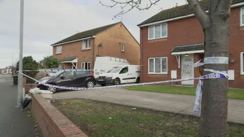 The image shows a residential street with two brick houses. There is a white van and a black car parked in the driveway of one house. The area is cordoned off with police tape that reads "POLICE NOTICE." The tape is wrapped around a tree and extends to the front of the house, indicating that this area is restricted. There are no visible people in the image.