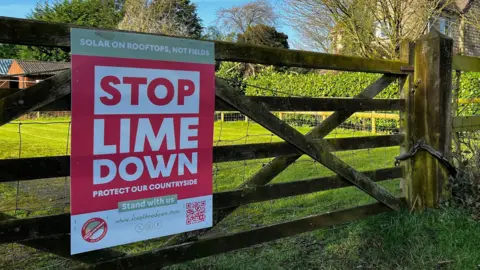 A red sign with white writing urging people to "Stop Lime Down" is fixed on a five bar wooden gate. Behind the gate is a grassy field, and a stone house.