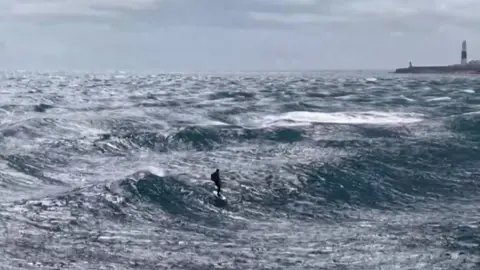 Emma Bridge A lone figure rides a hydrofoil above choppy, windswept waves, dwarfed by vast open sea as a lighthouse stands on the distant coastline under a grey sky.