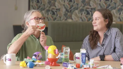 A screenshot of a video on the www.autismtoothcare.com website. Children's occupational therapist Saskia Grassie is demonstrating how to brush teeth using a toothbrush and a prop of a mouth. She's watched on by paediatric dentist Lauren Pickle. 