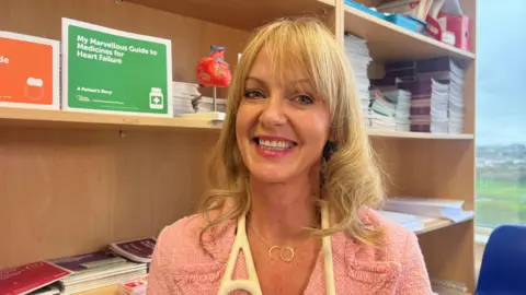 a woman with blonde hair and a pink jacket smiles at the camera. She is in front of some bookshelves and is wearing a stethoscope around her neck