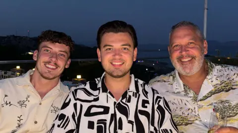 The Famileigh Travis, Harrison and Paul, all smiling, looking at the camera. Travis has a cream and black patterned shirt on, Harrison has a black and white shirt on and Paul, has a floral shirt on and is holding a glass.