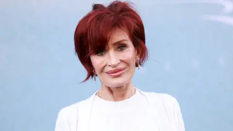 Getty Images A woman with short red hair and a white top in front of a pale blue background