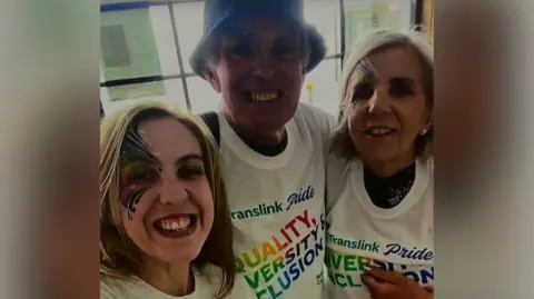 Family handout Natalie McNally smiling at the camera with her parents Noel and Bernie McNally. All three are wearing white Translink Pride t-shirts with a rainbow coloured logo which reads: "Equality, Diversity, Inclusion". 