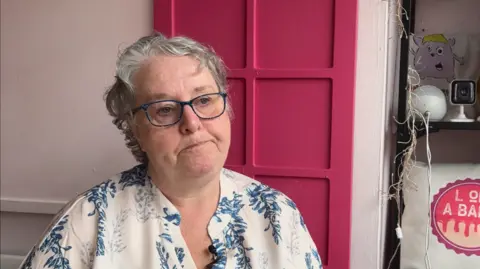 Lindsey Brown is wearing a white shirt with a blue floral pattern. She has short grey, curly hair and is wearing dark blue framed glasses. She is pictured seated in a cafe with pink walls. 