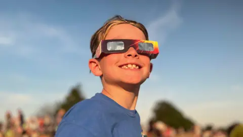 Nicola Rees/BBC A boy in a blue T shirt wearing solar glasses grins
