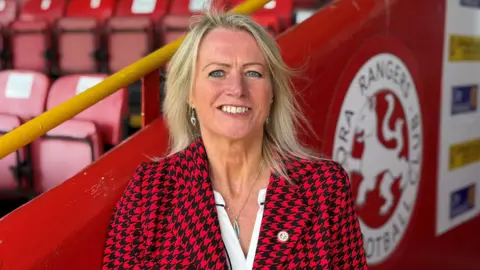 Mary Stewart has blonde hair and is wearing a red and black patterned jacket over a white top. She is smiling as she stands outside at Brora Rangers' ground.
