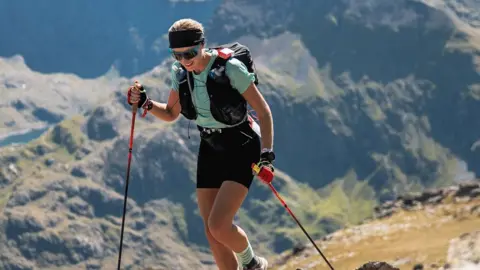 A woman with walking poles and a backpack is trekking across mountain terrain. She is wearing sunglasses and a headbad, with a green tshirt and black shorts. 