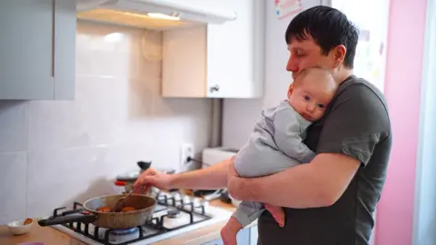 Stock photo shows a person holding a baby stirs food in a frying pan on a gas hob in a kitchen