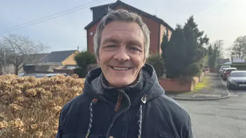 An image of Wayne Dixon, who is wearing a zipped up navy coat with blue and white drawstrings. Wayne is smiling with grey hair. He is standing on a street in front of a bush, with a row of terraced houses in the background.