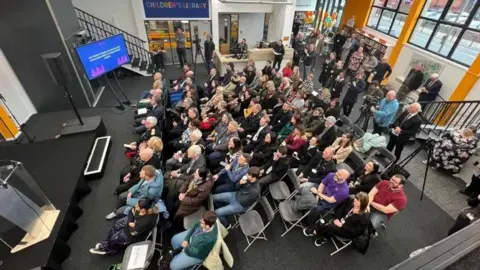 An aerial view of a large group sitting on fold up chairs facing a small stage inside the library.
