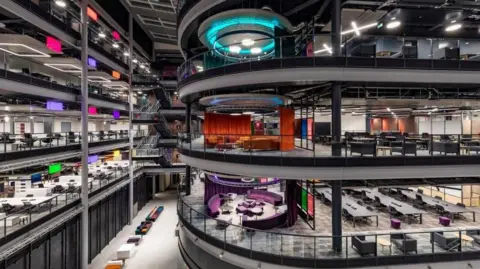 A colourful picture of the inside of BBC Wales' Cardiff headquarters. Three storeys of building can be seen, including stairwells, open plan offices and meeting areas.