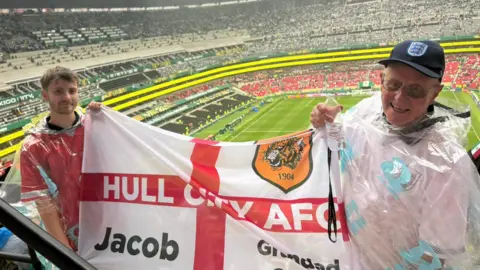 Jacob Allmendinger and his grandad, Geoff Golliker, wearing transparent rain ponchos hold a Hull City AFC St George’s flag in the stands of a large football stadium, overlooking the pitch before a match.