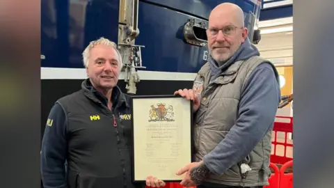 Paul Watling who is standing on the left, is presenting Richard Kirk, who is standing on the right, with a certificate. Both men are wearing gilet jackets and are looking directly at the camera and are smiling.