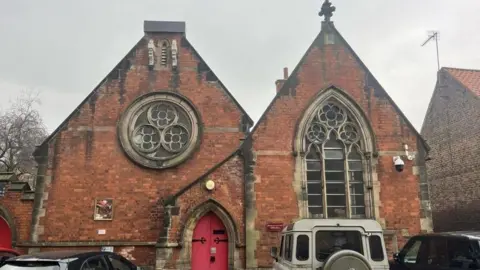 Beverley Minster Parish Hall, with its bell tower missing. There are cars parked in front of the red brick building.