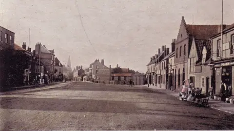 March & District Museum Black and white photo of March Broad Street from around 1900.
Shops either side of a wide road, a church steeple can be seen on the far left-hand side, there is no traffic at all, not even horses or carts, there are a few people that can be seen in Victorian clothing.