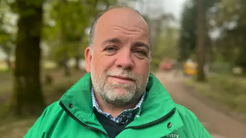 Richard Burke is wearing a green Forestry England branded jacket with a blue jumper and a blue and white checked shirt. He has short grey hair and a grey beard and is looking at the camera. The background is out of focus but shows resurfacing works with diggers in the forest. 