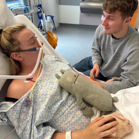 Katie Bain Katie is in the hospital bed with tubes attached to her nose, her partner Ben is tinning at her bedside and holding her hand.