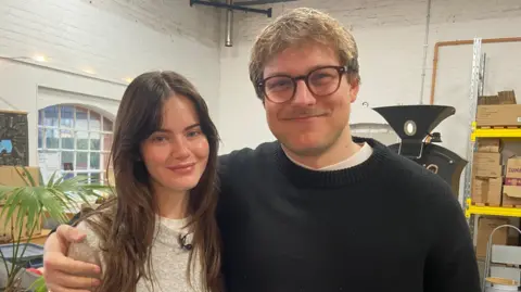 A man with blonde hair and glasses with his arm around a woman with brown hair. They are standing in a coffee roastery.