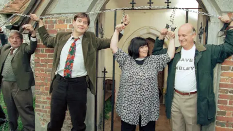 In this shot from an episode titled Celebrity Vicar, Geraldine Grainger, Frank Pickle, Jim Trott, and David Horton are depicted as characters involved in a protest outside the church. Geraldine and David are smiling 