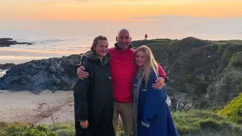 A man and two girls stand on the side of a cliff, which overlooks the beach and the sea.