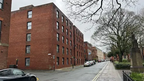 Edwin Williams Winckley Square and Cross Street in Preston as it is today. A large five story, plain, square red brick building replaces the stone one with an asphalt road with parked cars and trees to the right, bare of leaves