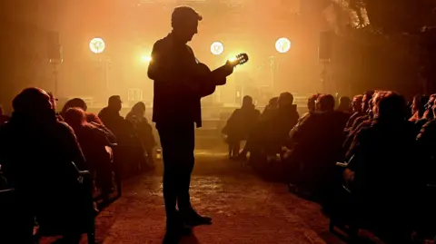 The image shows a silhouetted person standing in the centre of a narrow venue or tunnel-like space, holding and playing a guitar while facing a small audience seated on either side. Bright stage lights glow behind the performer, creating a warm orange haze that highlights the crowd, the rough floor, and equipment in the background.