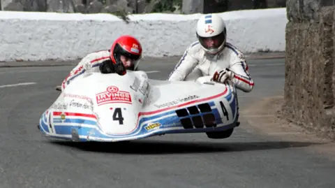 Southern 100 Archive Two men in white leathers racing a white and blue sidecar outfit on a road, with the driver wearing a red helmet