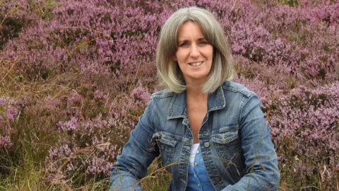 Supplied Heather Wainwright sitting in a patch of pink heather. She has blonde/white hair in a bob and is wearing a denim jacket. She is smiling at the camera.