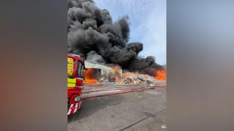 The front of a fire engine can be seen on the left of the image, with a large pile of cardboard on fire and large plumes of smoke billowing into the sky. A single-storey building can also be seen ablaze.
