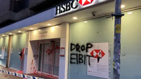 David Freezer/BBC Branch of HSBC bank with red paint splashed on the glass and doors. There is a large hole in one of the window panes, a smash mark on another, and lettering "DROP EIBIT" painted on a pane in black paint.