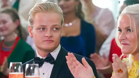 Marius Borg Høiby sitting next to his mother Norwegian Crown Princess Mette-Marit. He is wearing a black suit with a bow tie and his mother is wearing a yellow dress. Both have blonde hair.
