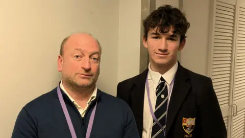 Aaron and Oliver standing next to each other. Aaron is wearing a blue jumper and Oliver is wearing a school uniform. They are both looking at the camera.