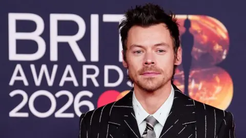 PA Media Harry Styles, who is wearing a black and white pinstriped suit with a tie and white shirt, is posing on the red carpet for the Brit Awards 2026 in Manchester. 