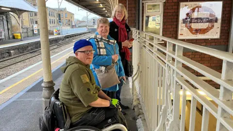 Ben, Pam and Anne are seen standing next to a railing on the train platform with the woven roundel on the wall next to them.