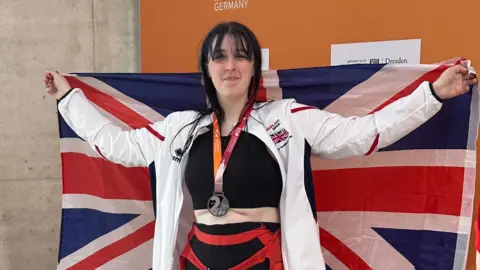 Supplied A woman with long dark hair, wearing a dark blue and red swimming costume is pictured holding a union jack behind her smiling at the camera